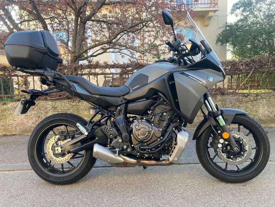 Extérieur urbain, moto touring Yamaha Tracer garée sur asphalte, environnement résidentiel avec végétation et bâtiments en arrière-plan