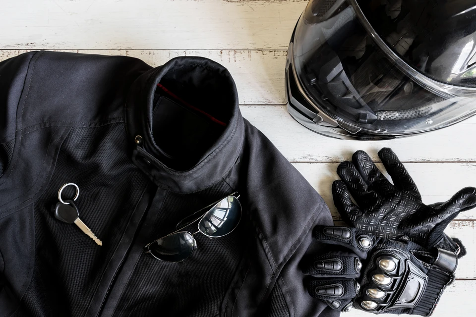 Équipement de motard disposé sur surface en bois: casque intégral noir brillant, veste de protection textile noire, gants de moto techniques avec coques de protection et lunettes de soleil
