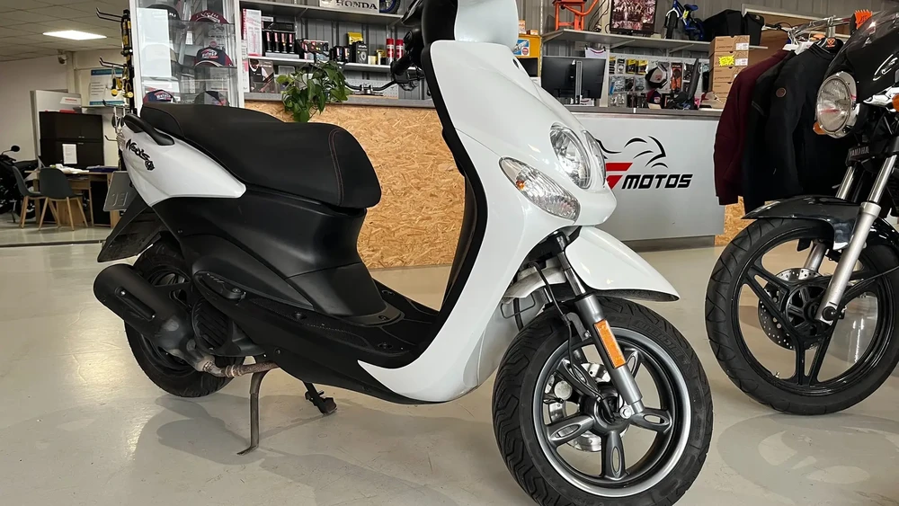 Intérieur d'un concessionnaire de motos avec scooter blanc et noir au premier plan, autres véhicules deux-roues à l'arrière