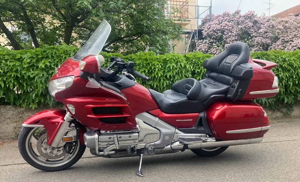 Scène extérieure urbaine avec une moto de tourisme rouge stationnée sur asphalte, végétation dense en arrière-plan avec arbres en fleurs et bâtiments résidentiels